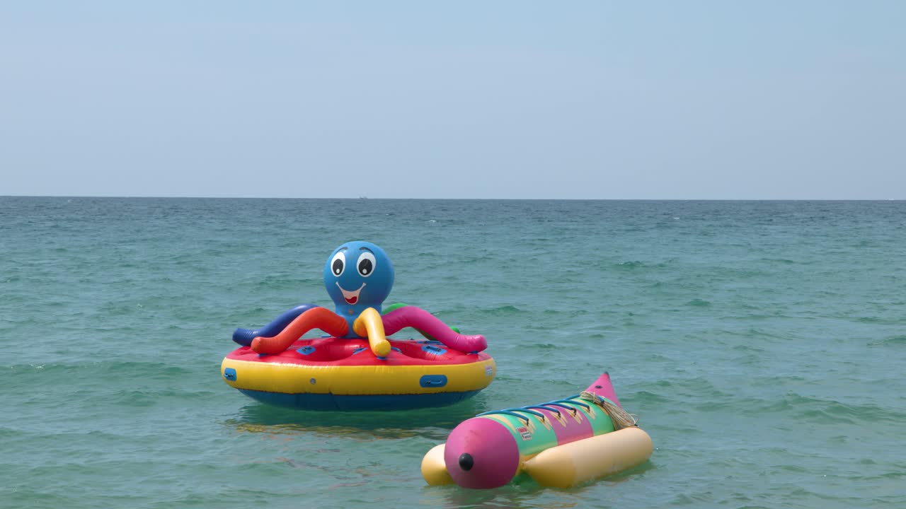 Static wide shot of vibrant inflatable water toys floating on calm turquoise ocean water