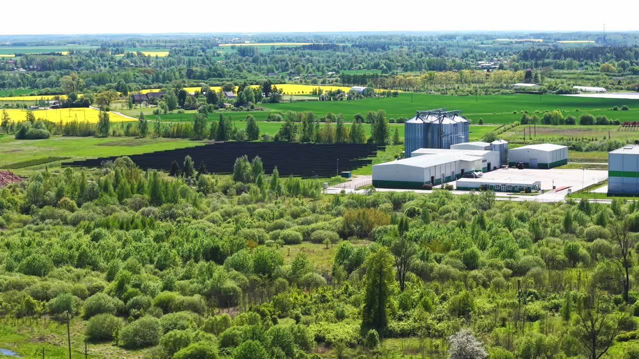 Aerial drone pass over industrial solar panel site in green terrain, efficient energy field next to silos and warehouses