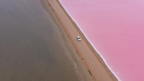 Excelente-Toma-Aérea-De-Un-Auto-Conduciendo-Por-Una-Carretera-Que-Divide-El-Lago-Macdonnell-En-La-Península-De-Eyre,-Sur-De-Australia,-Con-Agua-Marrón-En-Un-Lado-Y-Rosa-En-El-Otro