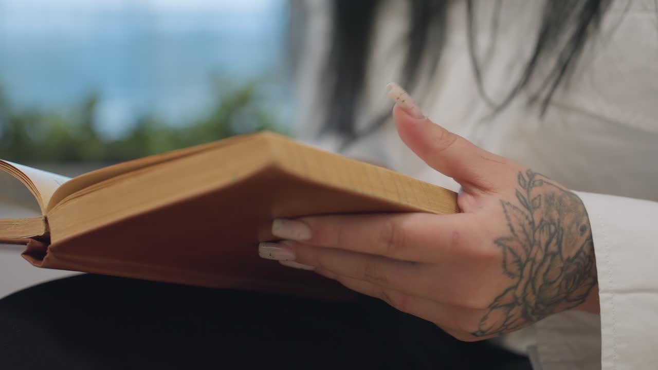 Close up of tattooed hand wearing white shirt gently flipping book page over open surface with soft blur of modern mall interior and hanging lights capturing rhythmic motion including glass railing
