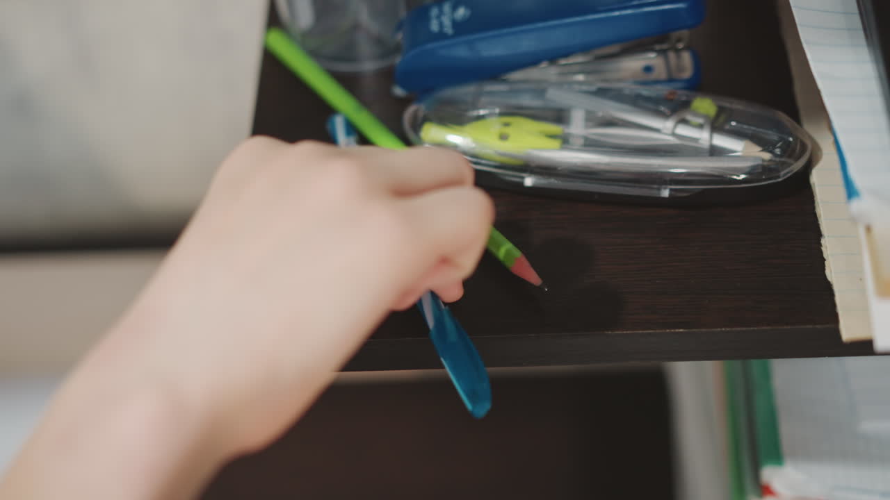 Grandson picks up fallen pencil from shelf and resumes drawing, small hand returns to sketching with green pencil, supplies and case visible, calm productive study and art moment