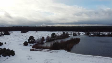 Aerial-view-over-snow-covered-beach-next-to-a-lake-inside-a-belgian-forest,-Lommelse-Sahara