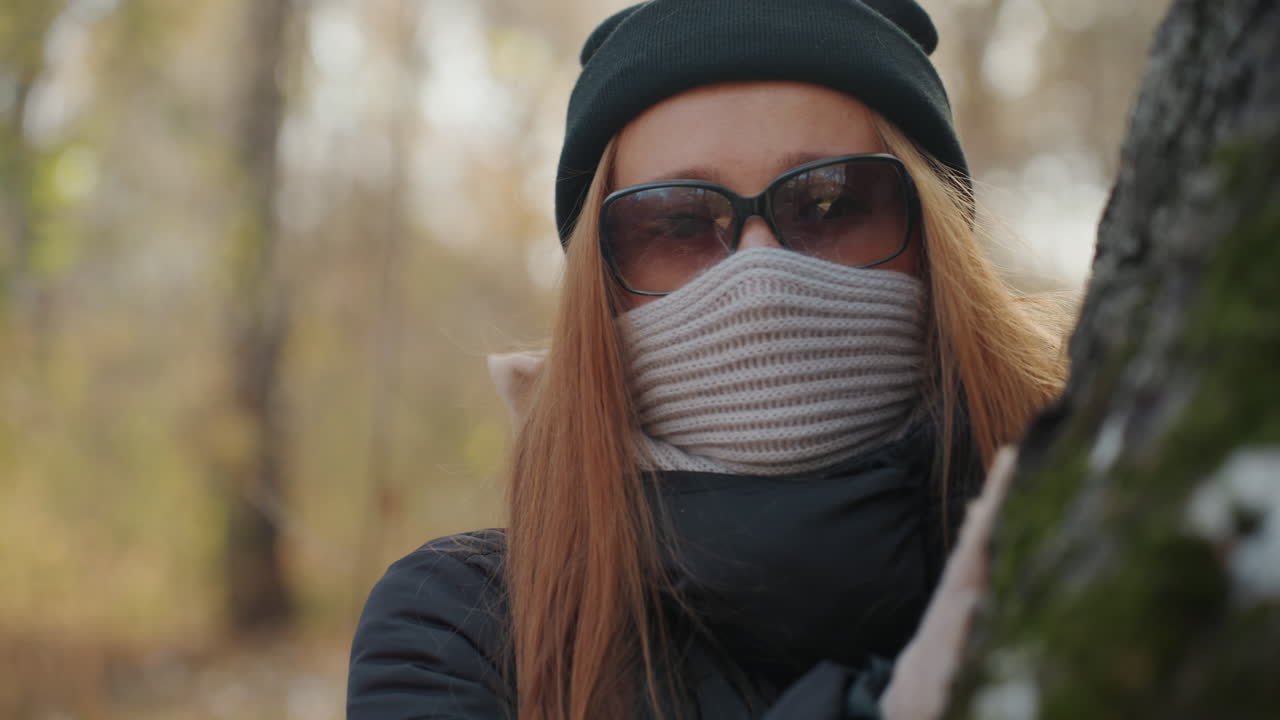 En maskerad tjuv kikar fram bakom ett träd i skogen. En närbild avslöjar en stickad halsduk och solglasögon som delvis döljer ansiktet, medan mössan och vinterkappan ramar in det röda håret. Mjuk snö ligger på trädets bark, och bilden har ett kort skärpedjup.