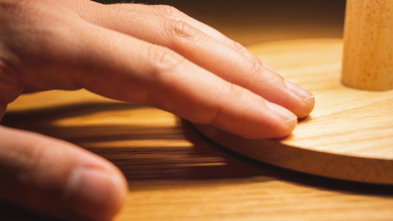Fingers coming left, hand sliding along wood base on tabletop, feeling grain with dowel, warm light