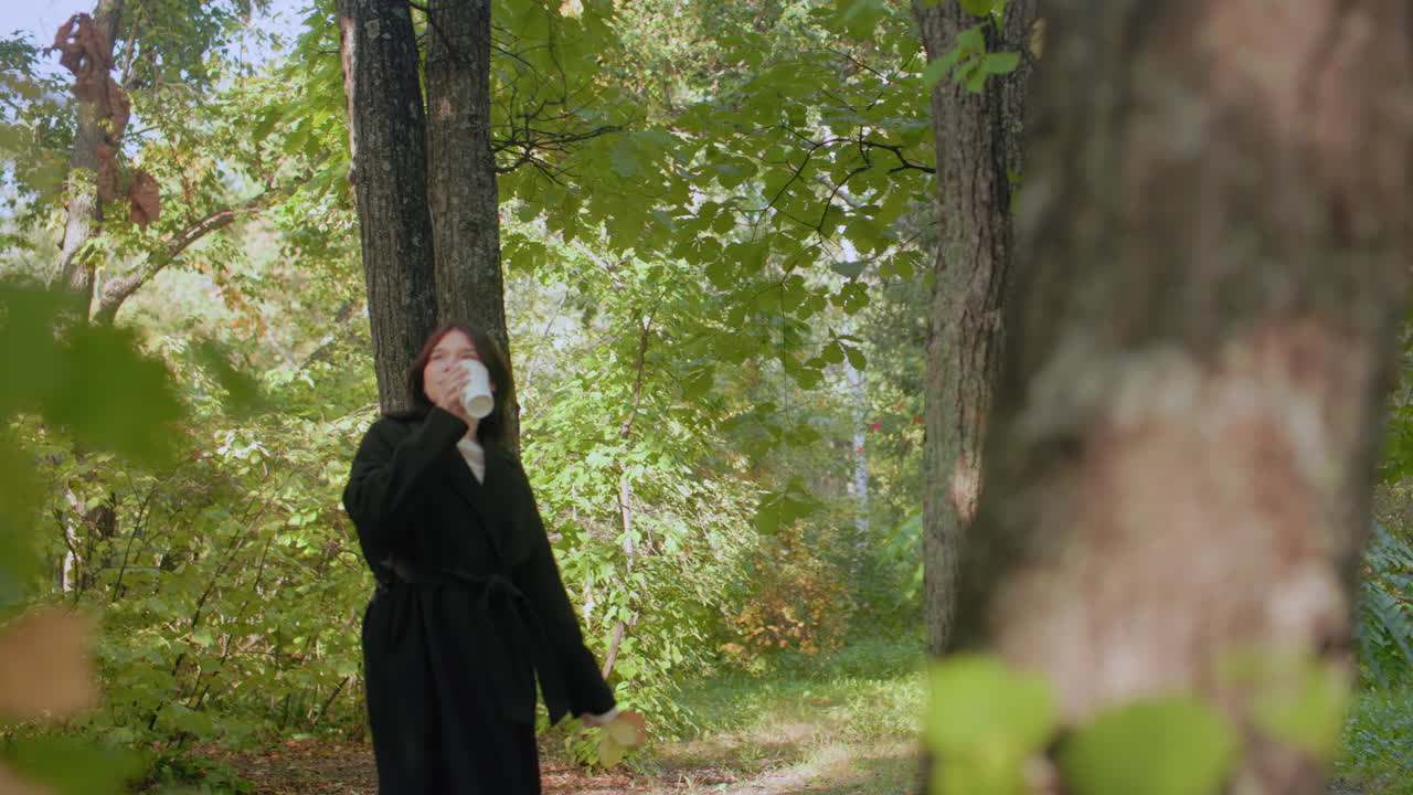 숲 속을 거닐며 따뜻한 차를 마시는 아웃도어 마니아, 햇볕 아래 눈을 감고 느긋하게 낙엽이 덮인 숲을 걸으며 고요한 가을 아침, 나무 사이에서 조용히 자연을 만끽해요.