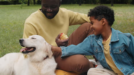 Alegre-Niño-Africano-Acariciando-A-Un-Perro-En-Un-Picnic-Familiar-En-El-Parque