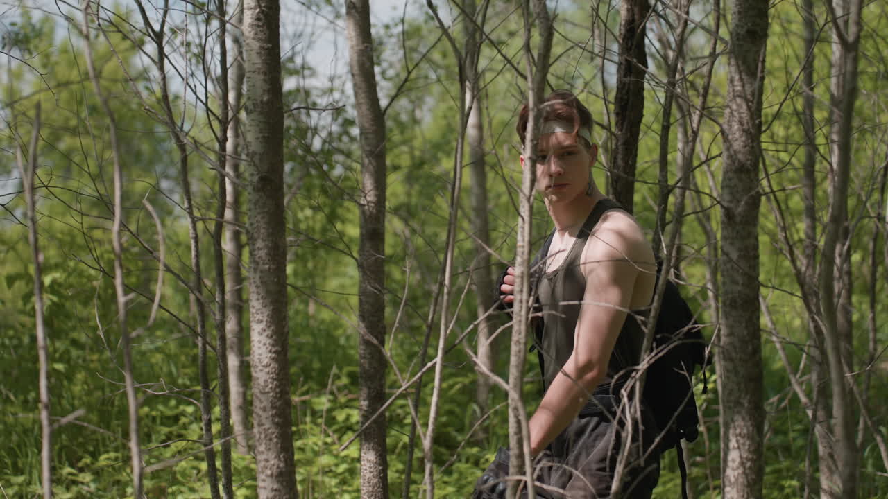 side view of warrior walking through narrow grove of thin tree trunks while scanning forest in particular direction sunbeams filtering through foliage onto fair skin and backpack strap held tight to shoulder