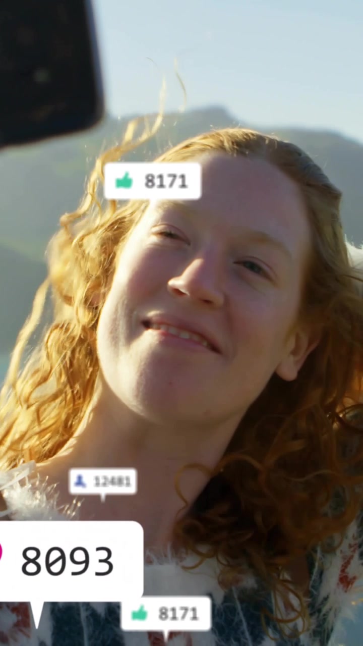 Mujer posando para redes sociales, cepillándose el pelo, sonriendo en la orilla, con un teléfono que aparece causando 'me gusta' animados.