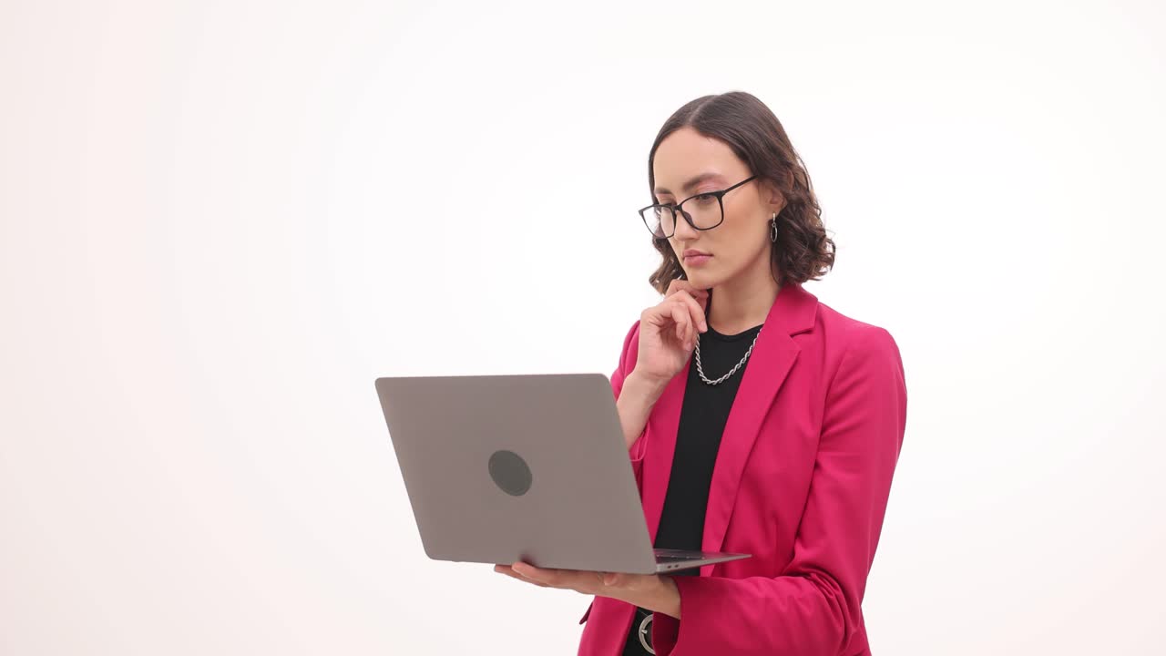 A woman in a pink blazer and glasses works on a laptop, appearing to concentrate or think