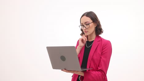 a woman in a pink blazer and glasses works on a laptop, appearing to concentrate or think