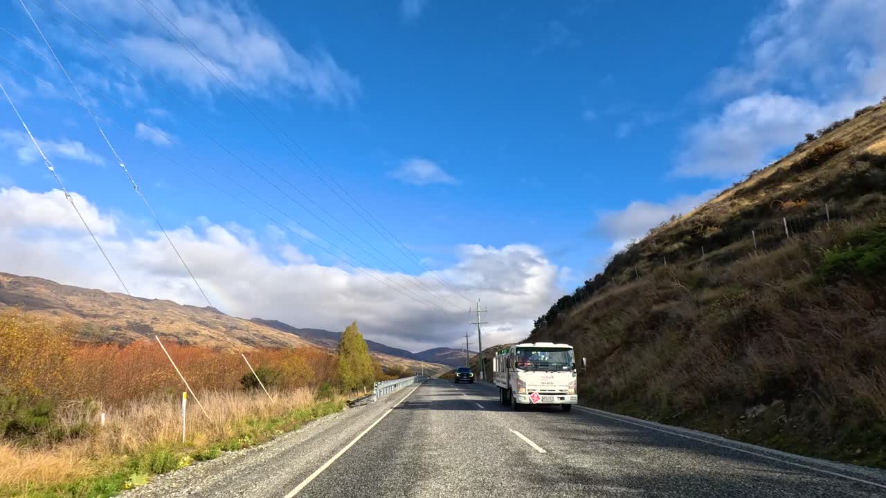 A vehicle travels a scenic road in Wanaka, New Zealand, surrounded by hills and clear skies, captured in bright daylight