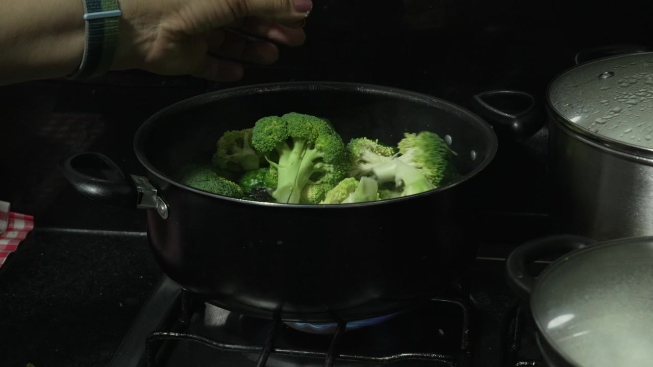Una mano lascia cadere cimette di broccoli verdi in una pentola d'acqua fumante su un fornello a gas in una cucina domestica buia, utile per video di cucina sana o nutrizione.
