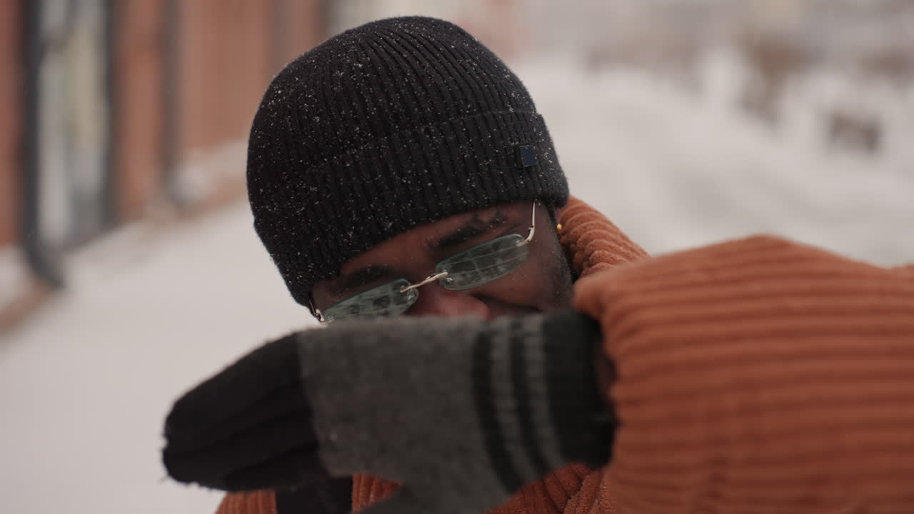 Uomo di colore con dolcevita arancione che si asciuga il viso, alle prese con il vento freddo e i fiocchi che cadono su un marciapiede urbano, primo piano spontaneo mostra occhiali, berretto, zaino ed espressione determinata durante l'inverno