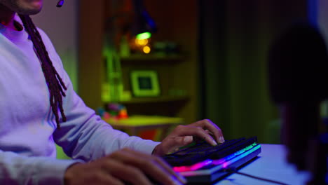 a young man with dreadlocks plays video games on his computer. he is wearing headphones and a microphone. the room is lit with colorful rgb lights.