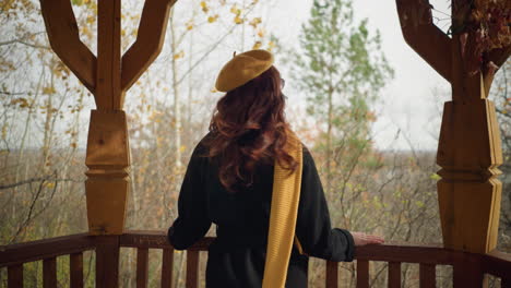 back view of young lady in yellow beret resting her hands on wooden rail, gazing into a forest as autumn leaves sway with the wind