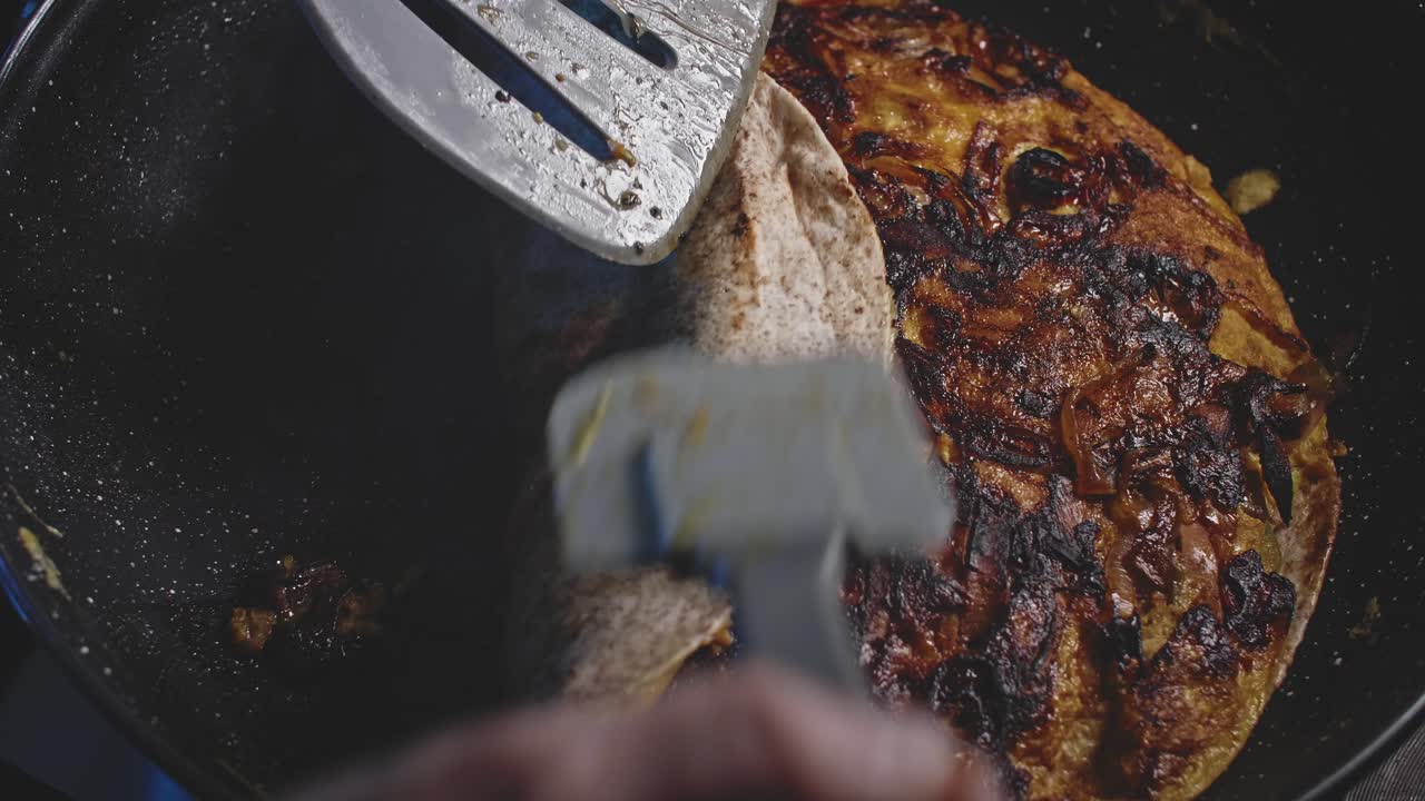 An overhead view looking into a skillet of a tortilla bed fried on top of egg, onion and bell pepper and a man uses a metal and a rubber spatula to roll it into a burrito