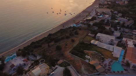 Toma-Aérea-De-La-Puesta-De-Sol-Naranja,-Sombrillas-Llenas-De-Playa,-Hoteles-Y-Pequeños-Botes-Flotantes-En-Grecia