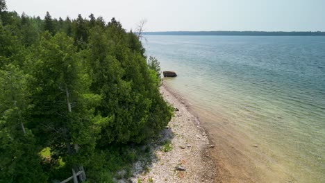 Aerial-view-descent-past-pine-trees-to-large-rock-on-beach,-Lake-Huron,-Michigan