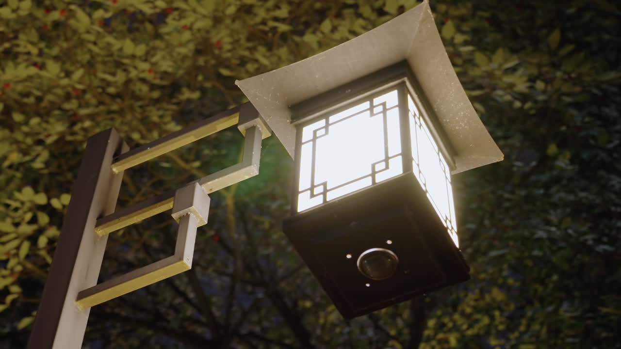 Gloeiende lantaarn hangt aan houten paal onder bladerdak in de schemering, Japans-geïnspireerd vierkant armatuur werpt warm zacht licht, metalen beugel en decoratief frame zichtbaar, zachte bokeh en schaduwspel