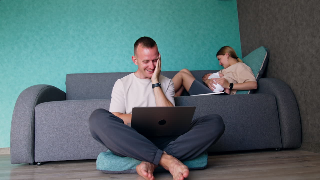 Caring blonde Caucasian woman nursing her tiny baby sitting on the sofa. Husbands sits on the floor with laptop in hands.