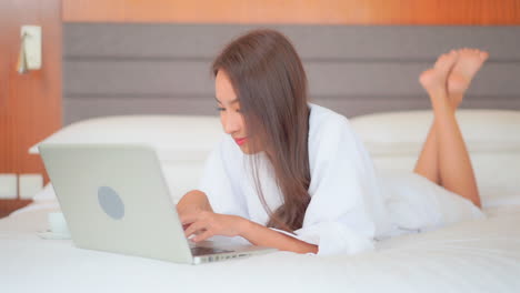 woman in bathrobe typing on laptop computer keyboard while lying on the bed in a luxury hotel