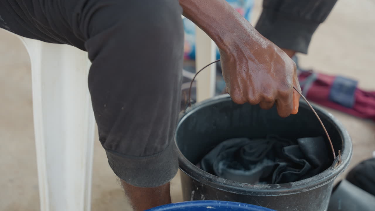 Mani di colore che lavano i vestiti in un secchio, scena in portico esterno, che strizzano un capo scuro con acqua saponata, seduto su una sedia di plastica, ritmo paziente, gesto di cura domestica, dettaglio tattile, luce solare soffusa.