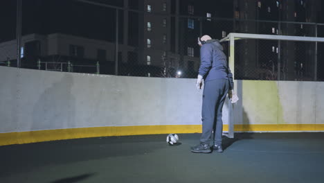 vista trasera de un hombre en ropa deportiva levantando hábilmente la pelota y pateándola contra la pared cerca del poste de la portería en la cancha al aire libre por la noche con edificios residenciales iluminados y entorno urbano en el fondo