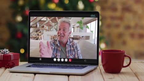 hombre caucásico de alto nivel saludando en una videollamada en una computadora portátil, con decoraciones de navidad y árbol