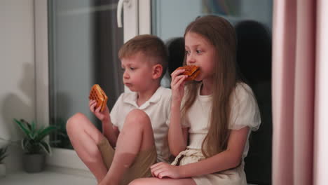 two cousins seated close to glass door relaxing while eating cookies and chatting warmly indoors, plants placed against wall nearby, evening atmosphere creating cozy backdrop