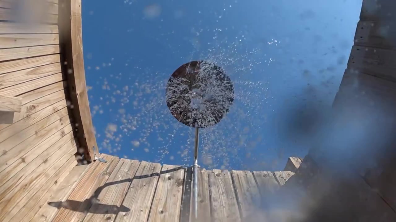 Doccia esterna con acqua fresca che spruzza dalla doccetta contro un cielo azzurro limpido e una recinzione in legno, offrendo un rinfresco estivo.