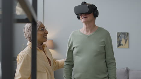 senior man using virtual reality glasses at home while an middle aged arabic woman helping him to find the right position in the room