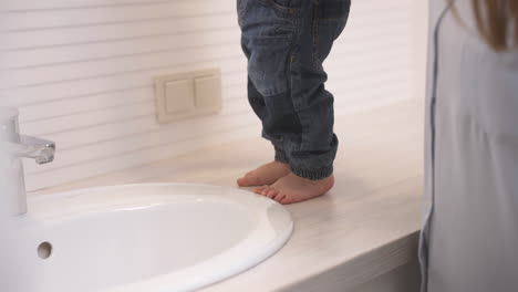 mother holds her baby over the sink in the bathroom while the baby holding a toy