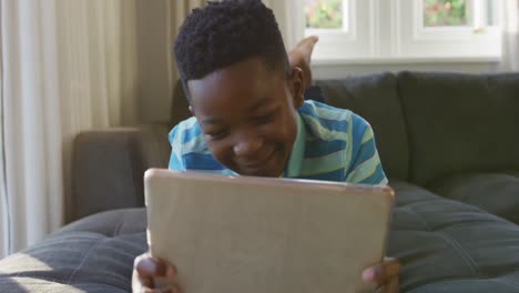 Glücklicher-Afroamerikanischer-Junge,-Der-Mit-Einem-Tablet-Auf-Der-Couch-Liegt-Und-Lächelt
