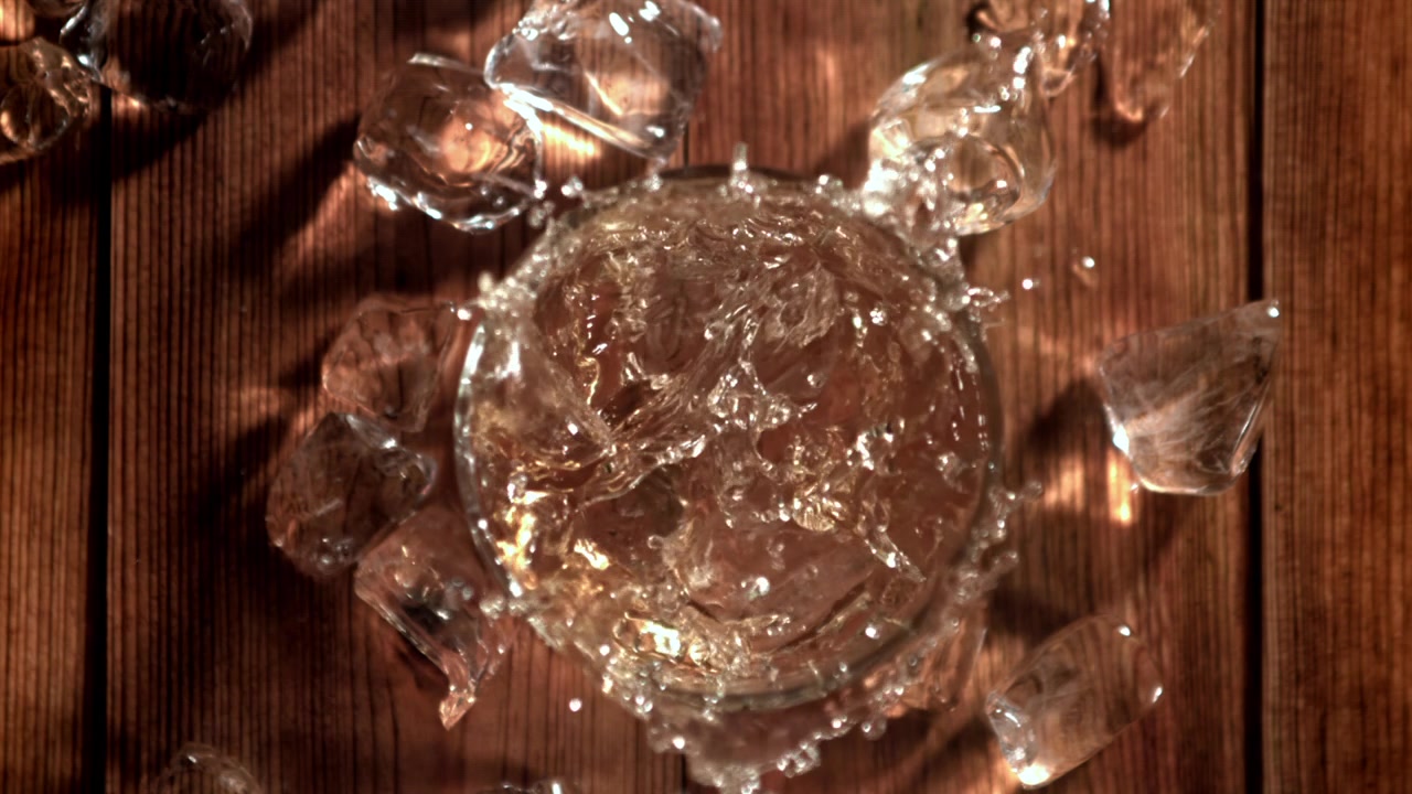 Cubetti di ghiaccio al rallentatore che si tuffano in un bicchiere di whisky su un tavolo di legno.