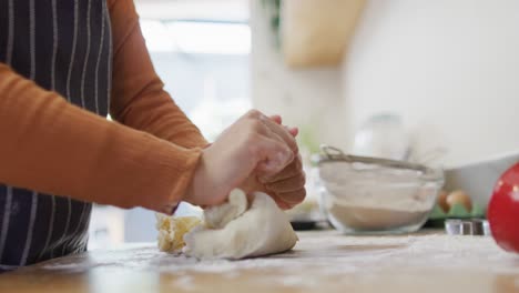 video of midsection of biracial woman baking in kitchen at home, kneading dough on worktop