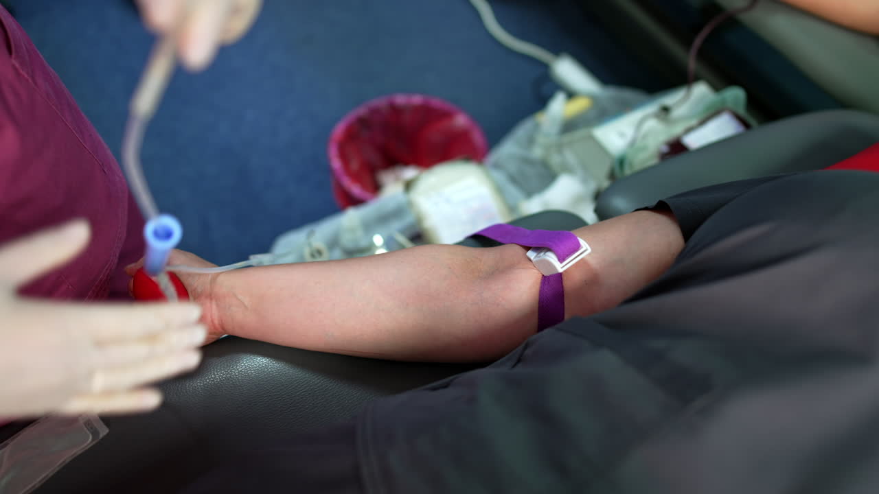 A person lies on the couch with a tourniquet on the forearm. A nurse prepares to attach the catheter to take some blood from the donor.