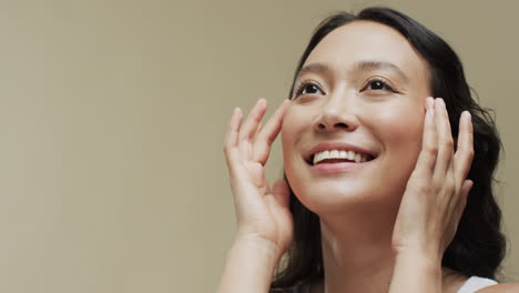 어두운 머리카락을 가진 행복한 아시아 여성이 복사 공간, 슬로우 모션과 함께 베이지 배경에 얼굴을 만지고 있습니다.