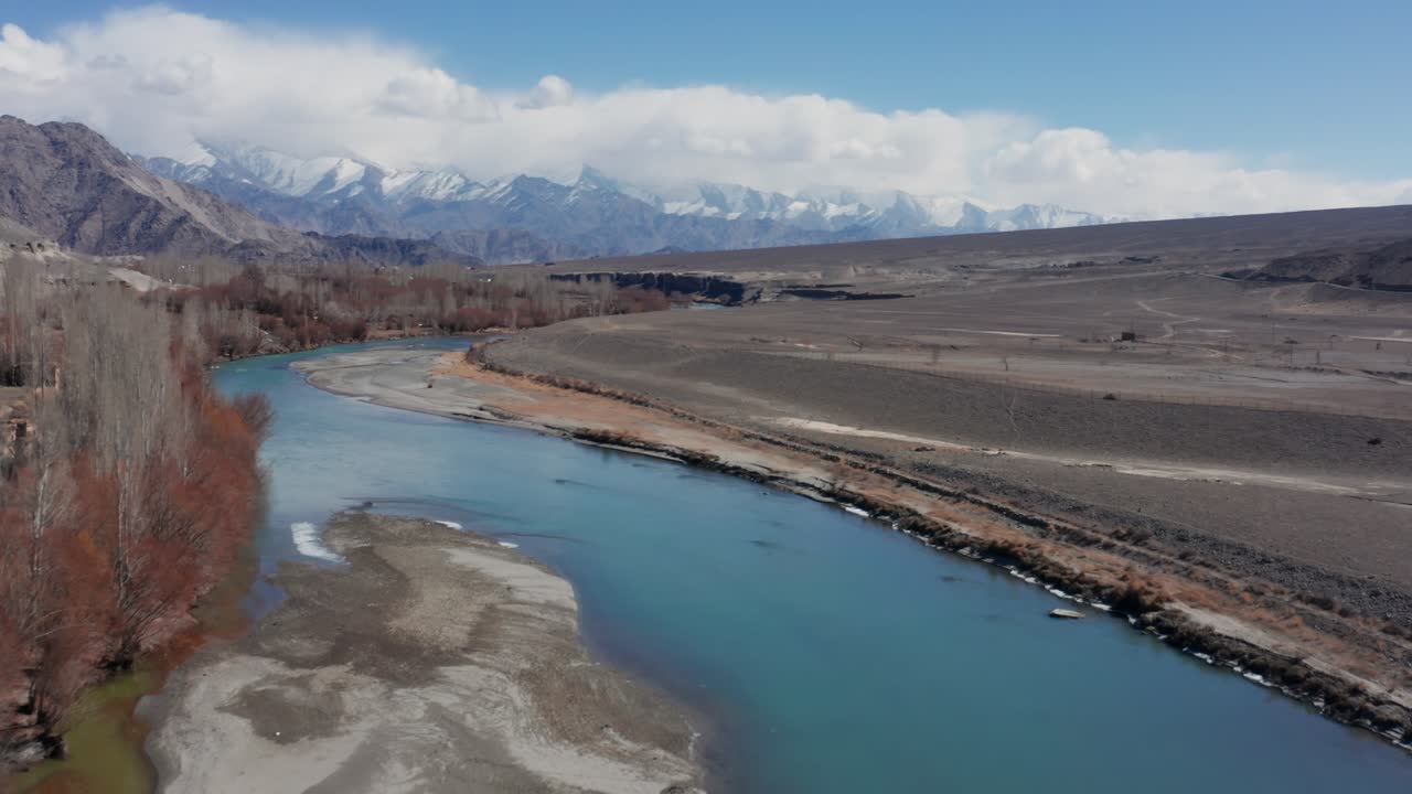 Vista aerea del fiume Indo turchese che scorre attraverso la valle invernale del Ladakh, delimitata da sponde di canneti marroni sotto cieli nuvolosi, con cime innevate in lontananza