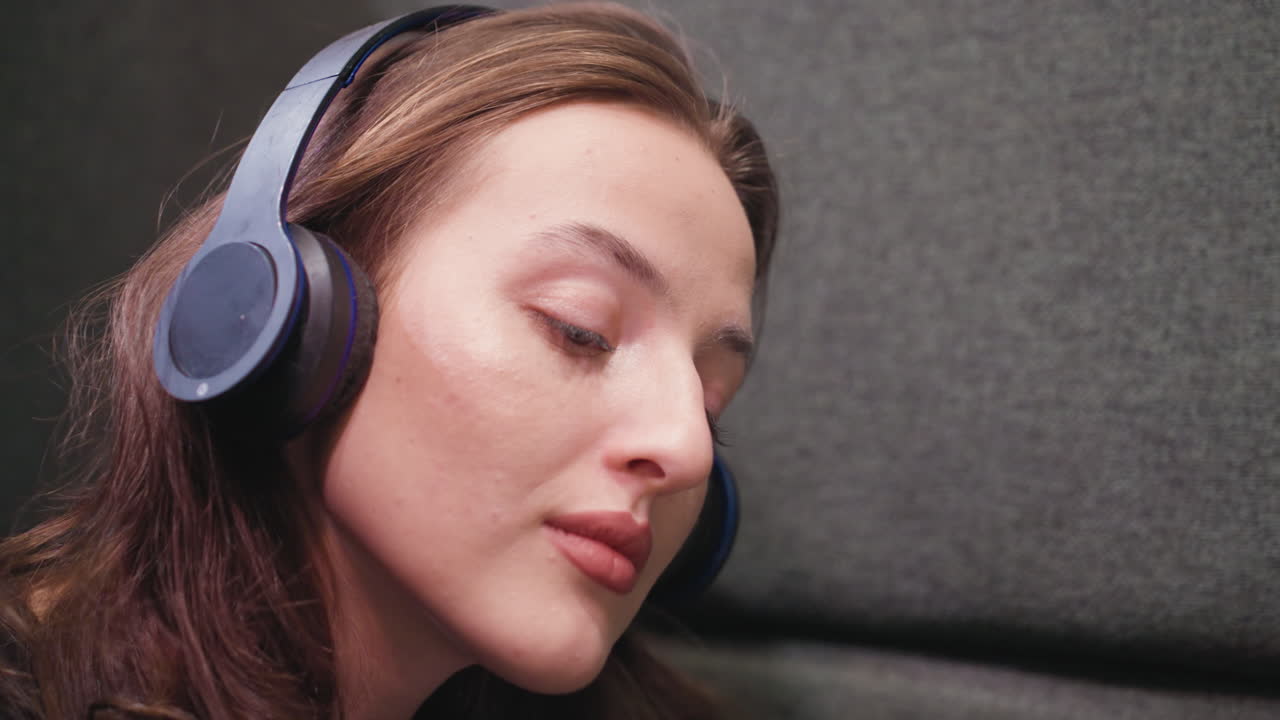Side close up of woman with soft features and brown hair wearing headset, gently nodding head with closed eyes. Calm expression reflects peaceful moment, likely immersed in music