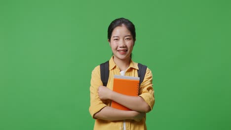 estudiante asiática con una mochila sosteniendo algunos libros y sonriendo a la cámara mientras está de pie en el estudio de fondo de la pantalla verde
