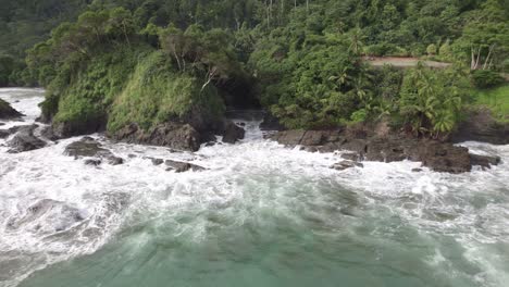 aerial dolly out rising on foamy turquoise sea near rocky shore and green forest hillside in dominicalito beach, costa rica