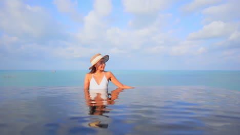 Fit-Woman-Model-in-White-Swimming-Suit-Leaning-Arms-on-the-Edge-of-Infinity-Pool-on-Turquoise-Seascape-Background-in-Bali-on-a-Sunny-Day-Slow-Motion