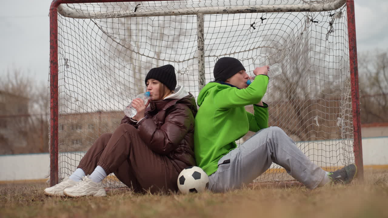 Два подростка отдыхают на футбольных воротах в шапках и куртках, сидя спиной к спине с футбольным мячом в ногах и пьют из бутылок в пасмурный день на городской площадке. Неформальный перерыв в тренировке показывает усталость