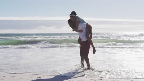 hombre afroamericano sonriendo y llevando a una mujer afroamericana a hombros en la playa