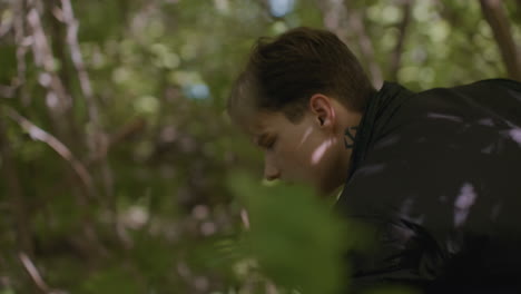 young tourist walking through forest shifts thin branch aside, then pauses briefly in motion to unzip backpack, surrounded by soft light and dense foliage as shadows dance across face and jacket