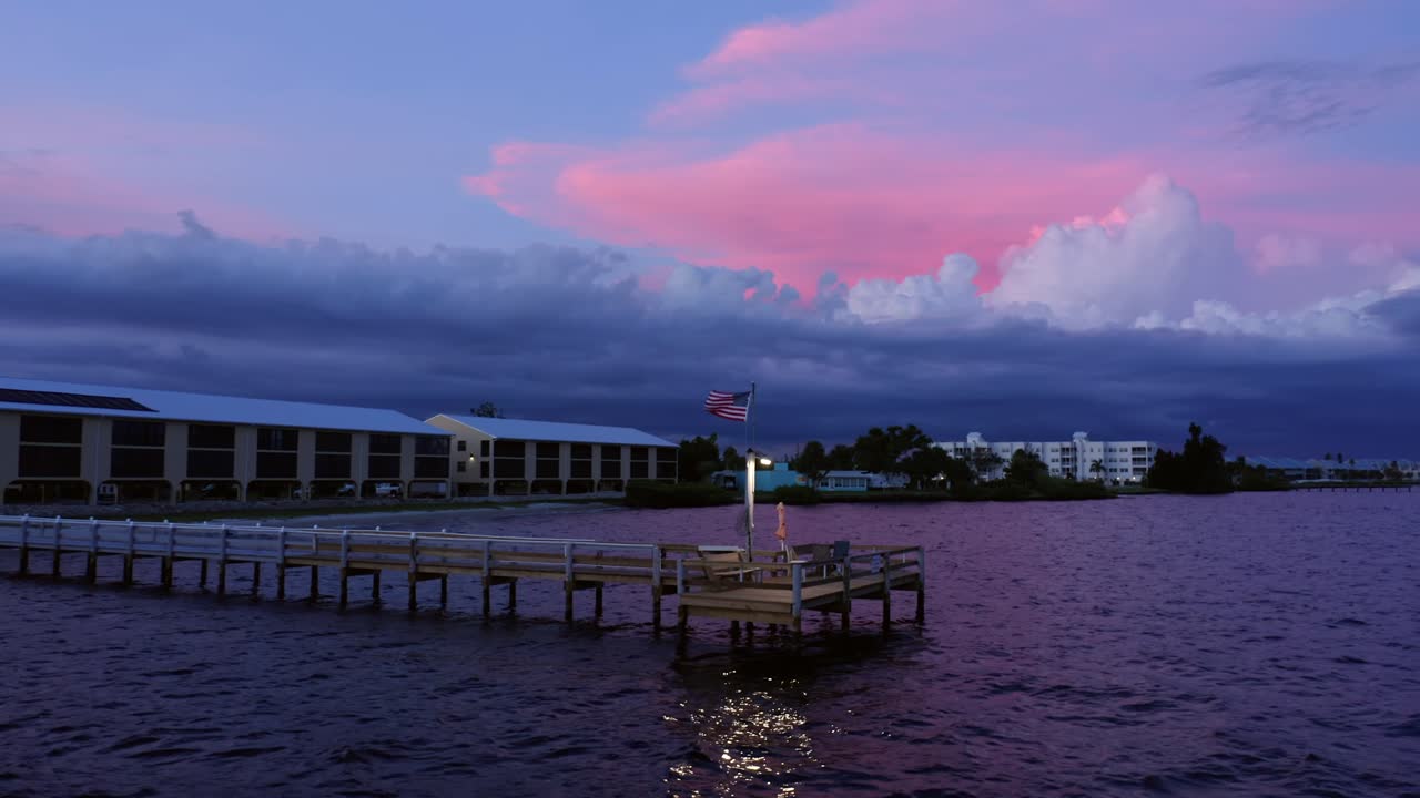A tranquil pier extends over rippling coastal water beneath a striking twilight sky as an American flag waves gently, reflecting serene evening atmosphere and vivid color contrast