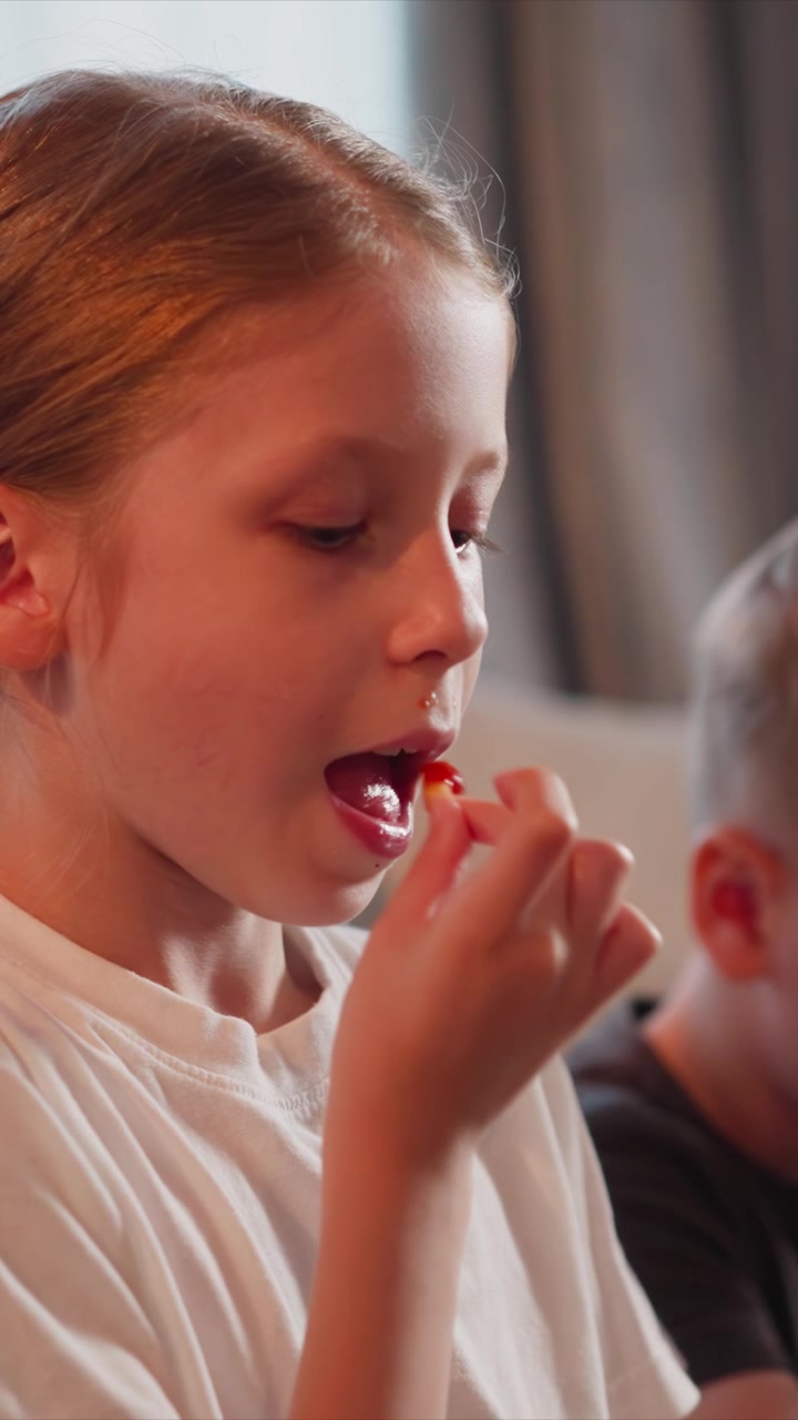 Primo piano di una bambina che mangia cibo mentre è seduta accanto a un altro bambino. La scena mostra dettagli della texture sulla sua maglietta bianca e un'illuminazione soffusa in un ambiente interno con elementi dello sfondo sfocati.