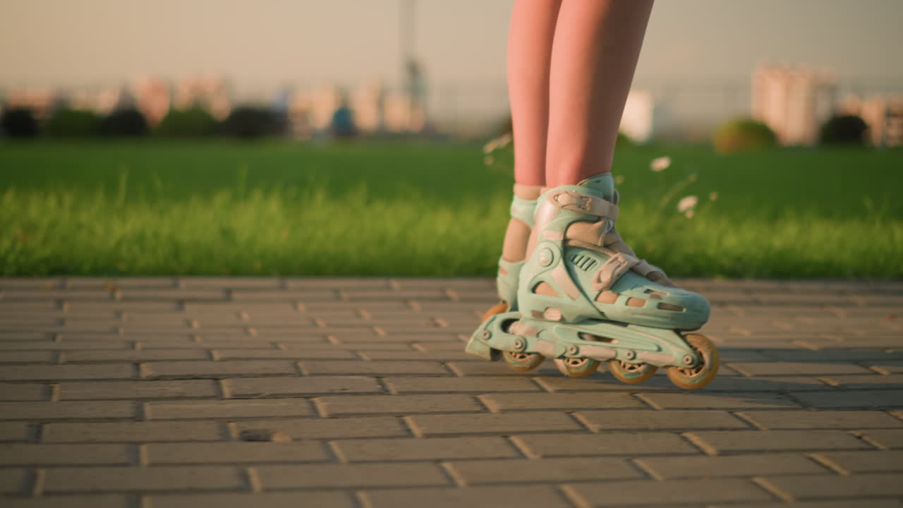 Leg view of person roller skating on interlocked path, rollerblades casting shadows on ground, next to grassy field, with blurred view of cityscape and buildings in background under evening sunlight