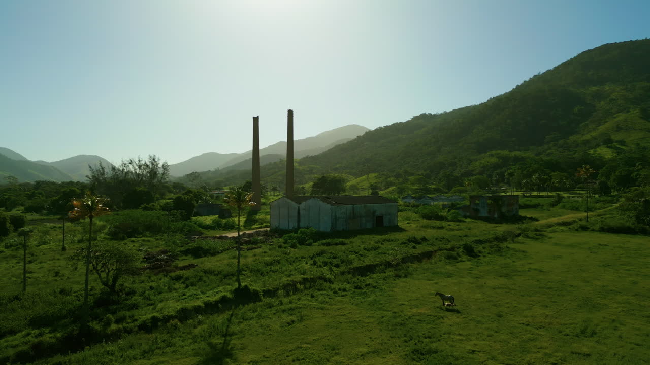 Drone terbang maju menuju pabrik gula dan etanol Santa Luiza yang terbengkalai di Sampaio Correia, Saquarema, Brasil. Ladang hijau dengan kuda yang merumput mengelilingi reruntuhan industri; bukit dan gunung