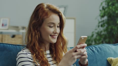 Young-Cheerful-Girl-With-Red-Hair-Tapping-And-Typing-On-The-Smartphone-While-Sitting-On-The-Couch-At-Home